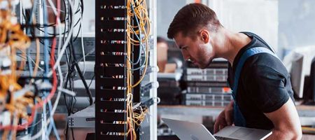 Junger Mann in Uniform und mit Laptop arbeitet mit Internetgeräten und Kabeln im Serverraum